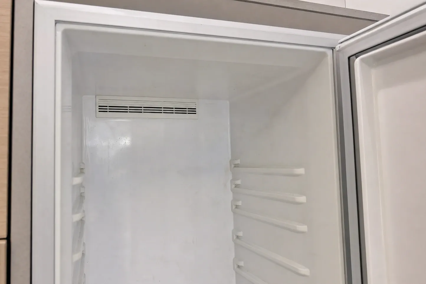 Empty white refrigerator column interior, door open, air vent in the rear wall and shelf rails on the door