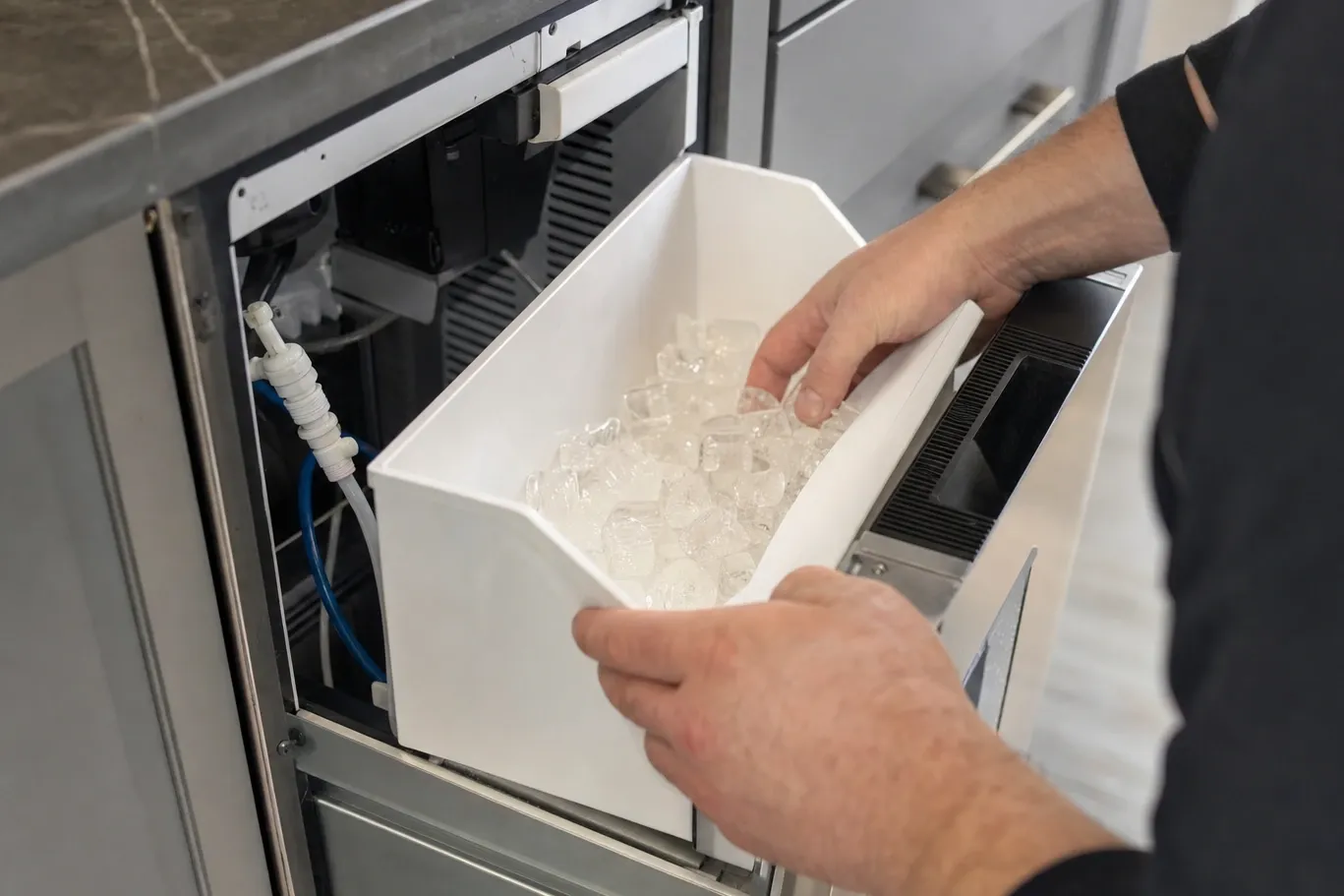 Hands lifting a bin of clear cubes out of an undercounter ice maker, water line and fittings behind